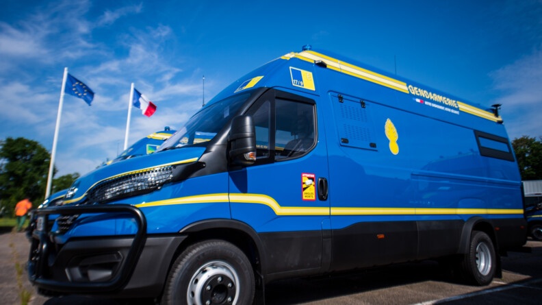 Véhicule de commandement et de transmission bleu avec des traits jaunes, stationné sur un parking. Derrière, au fond, les drapeaux français et européen.