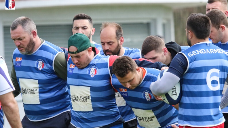 La première et deuxième ligne d'une équipe de rugby, au maillot bleu, se prépare à la mếlée. A droite, un joueur de dos tient le ballon sous son bras gauche
