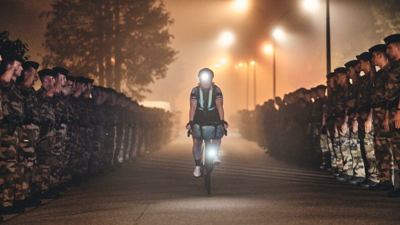 Une jeune femme cycliste remonte une allée de nuit, tout au long de laquelle se dresse une haie d'honneur de militaires en tenue. Les lamapadaires et la lampe frontale de la cycliste forment des points lumineux.