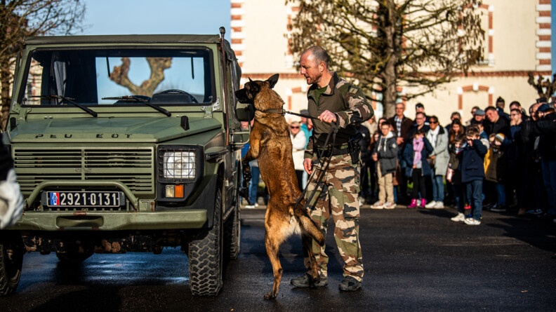 Une équipe cynophile fait une démonstration devant un public avec de nombreux enfants. Le chien vérifie une jeep, sur ses pattes arrière, tenu en laisse par le maître-chien.