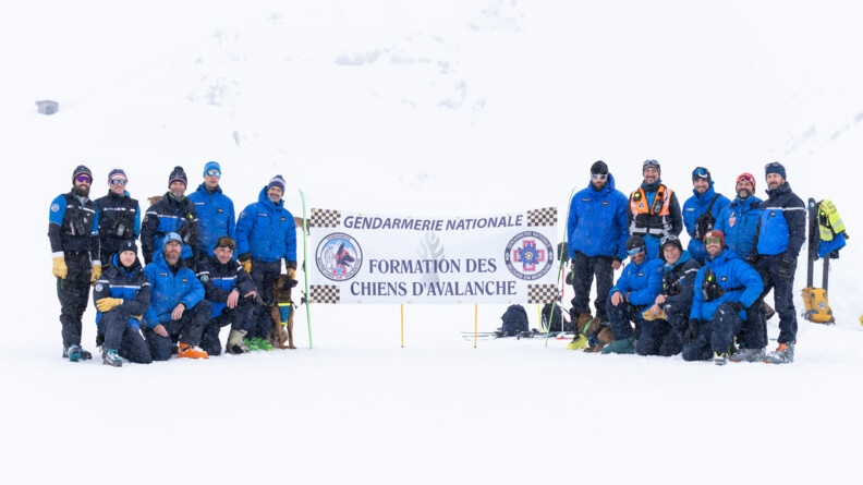 Un groupe d'hommes vêtus d'une tenue de ski bleue, se tiennent sur un sol enneigné, de part et d'autre d'une grande banderole portant l'inscription Gendarmerie nationale Formation des chiens d'avalanche.