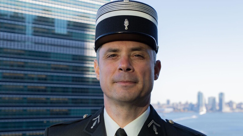 Portrait de face d'un colonel de gendarmerie, coiffé d'un képi, avec en arrière-plan les bâtiments new-yorkais et l'East-River