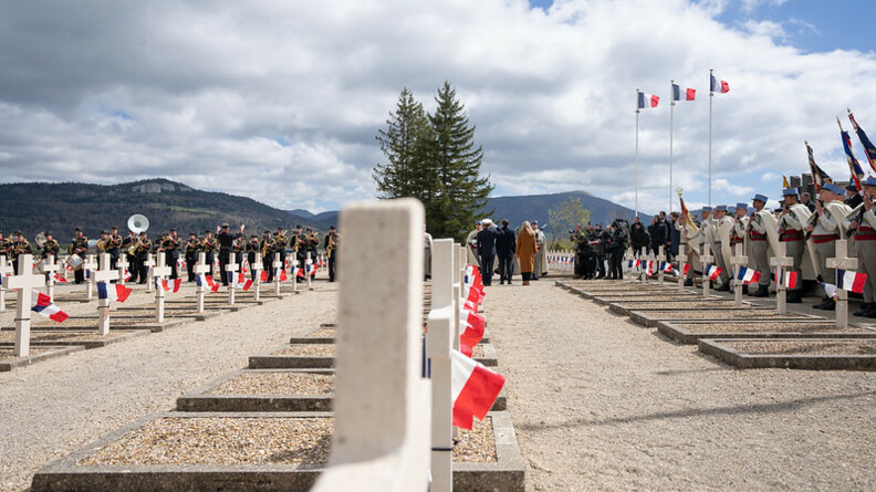 Image d'un cimetière aux croix blanches, sur lesquelles sont disposés des drapeaux français. Des militaires se tiennent debout en bordure des tombes.