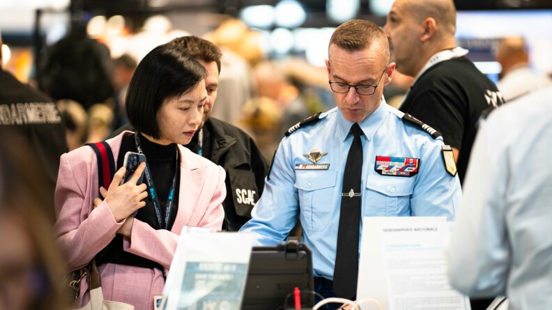 Devant une malette, un colonel de gendarmerie présente à une femme portant une veste rose un système de communication