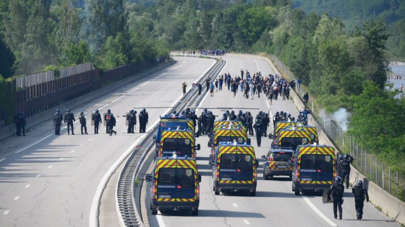 Au premier plan, une douzaine d'Irisbus vus de dos, sur l'A43. Devant eux, sur toute la largeur de l'autoroute, des gendarmes mobiles s'avancent vers un groupe de manifestants radicaux.