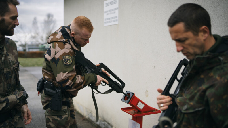 Un gendarme en treillis fait ses controles d'arme avec un FAMAS dans un tube à sable rouge