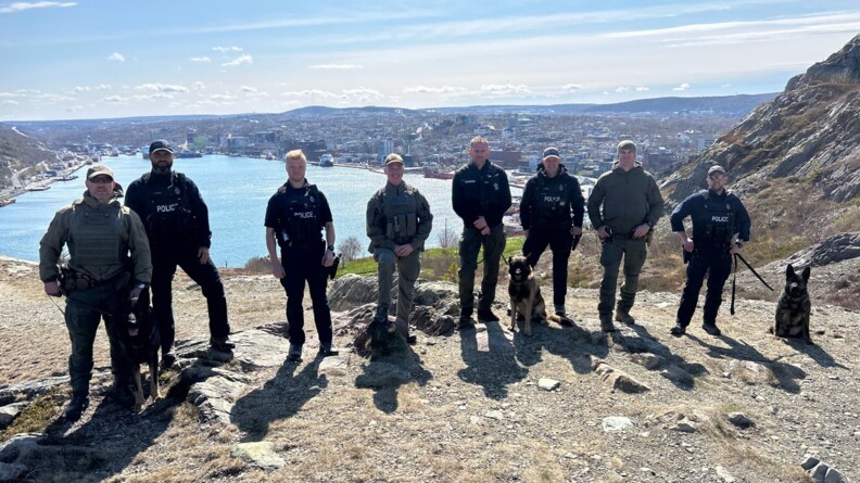 Posant sur une colline devant un paysage canadien (lac, ville, collines), 8 membres des polices canadiennes et de la gendarmerie française de Saint-Pierre-et-Miquelon avec leurs chiens malinois