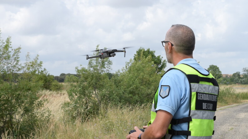 Un militaire de la gendarmerie de l'Air, à droite, pilote un drone en vol, au centre.