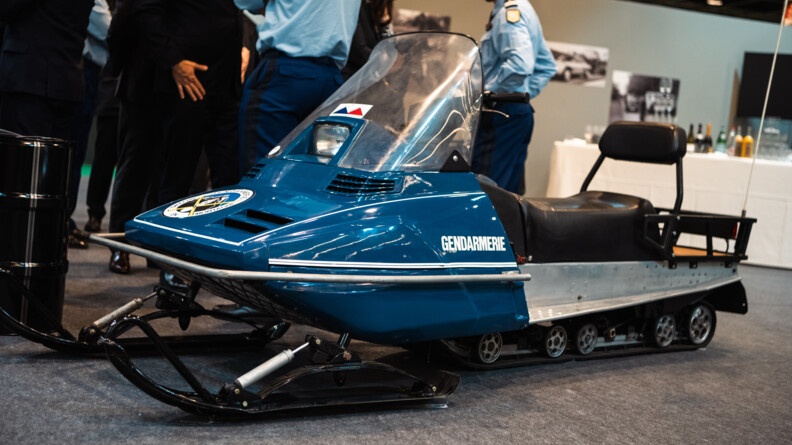 Photo d'une motoneige bleue utilisée lors des jeux olympiques d'Albertville en 1992 en premier plan. Au deuxième plan, des gendarmes discutent debout.