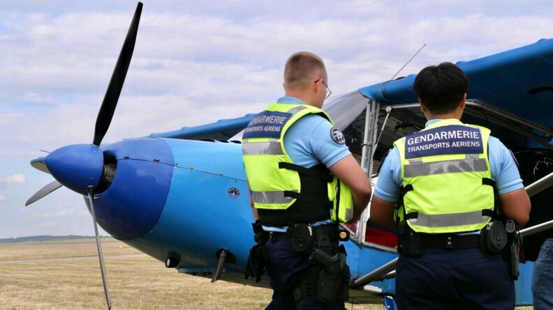 Des gendarmes de la GTA sont devant un avion bleu. Ils discutent avec son pilote.