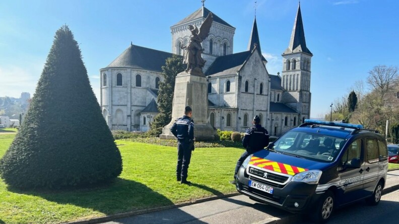 Deux gendarmes de dos sur l'herbe qui entoure un monument aux morts. En bas à droite un véhicule gendarmerie vue de trois-quart face. A gauche un arbre taillé en forme de cône. En arrière plan une église sous un ciel bleu