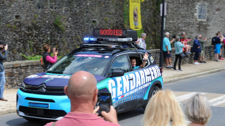 Un véhicule de la caravane gendarmerie du Tour de France avec les spectateurs qui filment sur les bas-côté. A la fenêtre arrière gauche du véhicule, une jeune gendarme souriante salue de la main