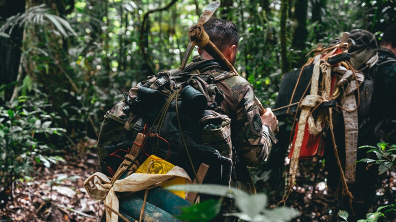 Un militaire marche en forêt afin d'atteindre un chantier illégal. Il porte une pioche sur son épaule.