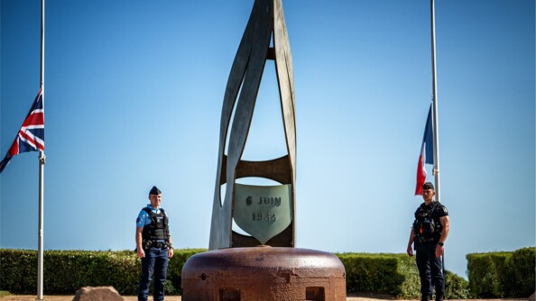 Deux gendarmes entourent le monument dédié aux morts du 6 juin 1944. De part et d'autre, les drapeaux français, à droite, et britannique, à gauche.