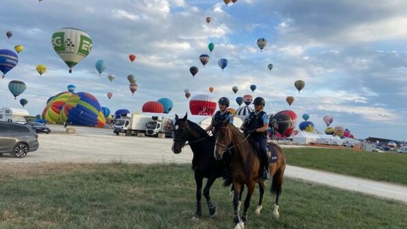 Deux cavaliers en patrouille équestre sur une zone herbeuse, à proximité d'un parking, avec un vol de montgolfières en fond.
