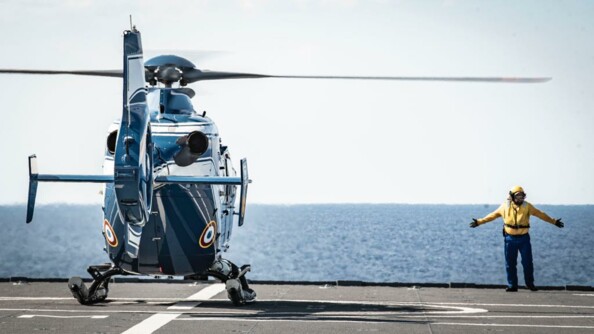 Un hélicoptère de la gendarmerie nationale en train de se poser sur le pont du porte hélicoptères Mistral de la Marine nationale, d'où un signaleur le guide. En arrière-plan, la mer.