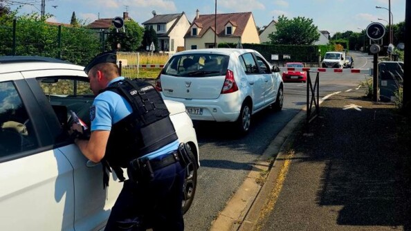 Un gendarme, vu de dos, dispense des conseils de prévention par la vitre du passager, à un conducteur arrêté derrière un autre véhicule, devant un passage à niveau (en arrière-plan), dont la barrière est baissée.