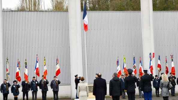 Six personnes, autorités civiles et militaires au salut, vues de dos, face au drapeau en train d'être hissé. De part et d'autre du mât des couleurs, une demi-douzaine de porte-drapeaux.