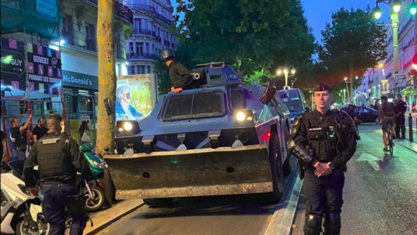Deux gendarmes mobiles orientent la progression d'un véhicule blindé en plein coeur de la ville. Un troisième gendarme apparait au niveau de la tourelle.
