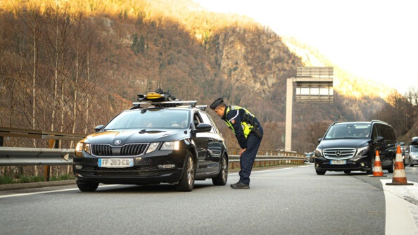 Un gendarme avec une chasuble jaune conseille un automobiliste sur la route. On peut voir des skis sur le toit du véhicule et de nombreuses voitures en arrière plan.