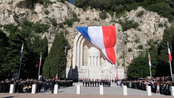 Sur une grande place, devant un majestueux monument aux morts est drapé d'un immense drapeau bleu, blanc, rouge, se tiennent des militaires et des civils. On aperçoit des arbres et une haute roche  contre laquelle le monument aux morts prend appui.