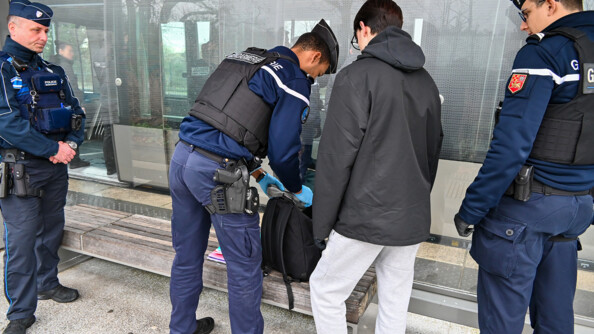3 gendarmes sont entrain de fouiller le sac à dos d'un jeune homme sous un abri de train.