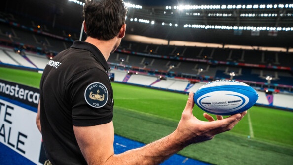 Gendarme EOR (Explosive ordonance reconnaissance) de profil tenant de la main droite un ballon de rugby aux couleurs de la gendarmerie devant la pelouse du Stade de France.