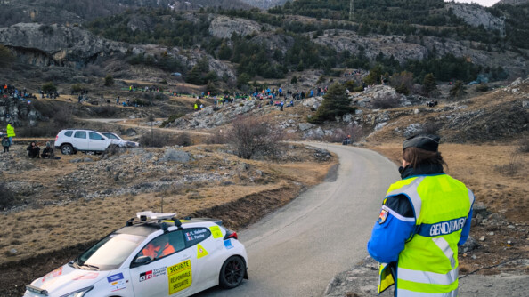 Dans les alpes du sud, des spectateurs regardent la course pendant que les gendarmes, debout sur un rocher, observent le bon déroulement du rallye.