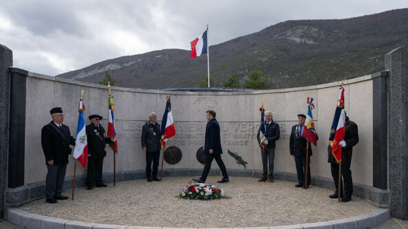 Devant un monument circulaire en pierre, le chef de l'Etat, Emmanuel Macron, marche devant des anciens combattants ou amis de la Résistance, se tenant debout, tenant le manche d'un drapeau dans leur main. Au sol est posée une gerbe de fleurs recouvrte d'une bannière bleue, blanc, rouge.