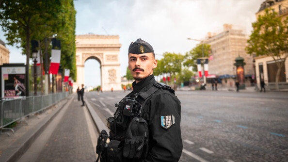 Un gendarme mobile au centre, de trois-quart face, regardant au loin, avec en arrière-plan les Champs-Elysées et l'Arc de Triomphe
