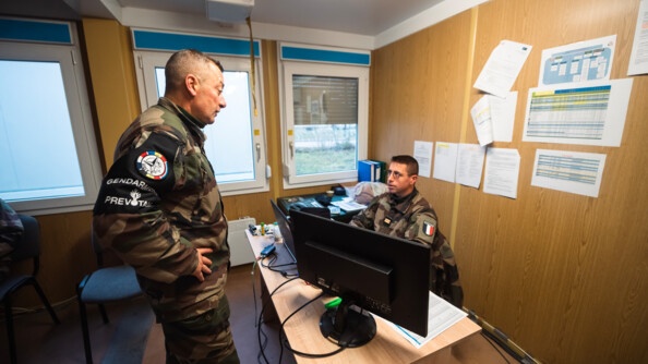 A gauche, de profil droit, les mains sur les hanches, un prévôt échange avec un lieutenant de l'armée, assis derrière son ordinateur