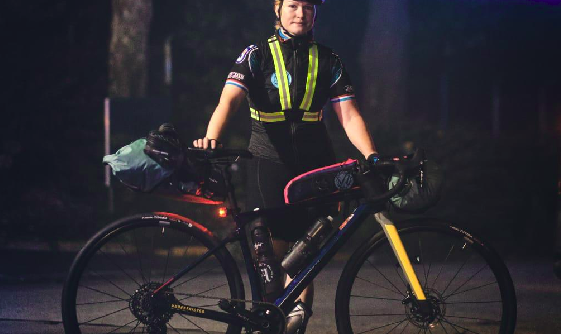 Une jeune femme cycliste pose de nuit devant son vélo. Elle se tient debout, une main posée sur la selle, l'autre sur le guidon de son vélo. Elle porte un casque et des lunettes par-dessus au niveau du front.