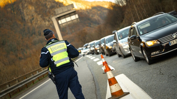 Un gendarme avec une chasuble jaune  vérifie l'état du trafic sur la route. A droite, on voit une colonne de voitures. Au fond, les montagnes.