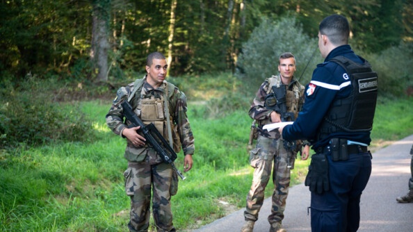 Deux militaires de l'armée de terre, armés chacun d'un fusil, sont debout sur la route  à la lisière d'une forêt. Ils échangent avec un gendarme que l'on voit ici de dos.