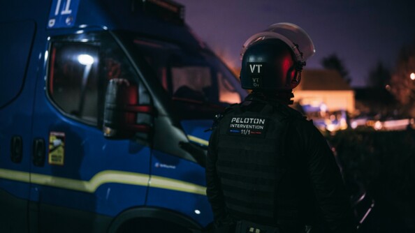 Un gendarme mobile casqué de dos; à sa gauche un véhcile de maintien de l'ordre. Au loin des bâtiments et véhicules gendarmerie à la lueur de l'aube