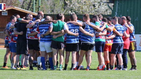 Environ 25 hommes en tenue de rugby, de différentes couleurs, en cercle, se tiennent par les épaules et le dos