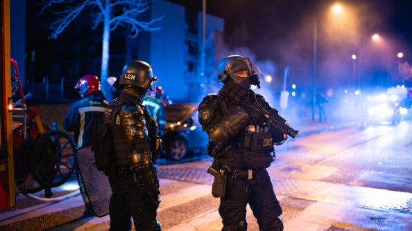 Dans la nuit, photo de deux gendarmes mobile dans la rue. En fond, des pompiers se préparent à éteindre un feu de véhicule pendant que les gendarmes sécurisent la zone.