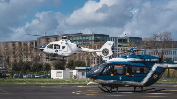 Hélicoptère bleu de la gendarmerie au premier plan, encore au sol, et hélicoptère blanc du SAMU au second plan, en phase de décollage.
