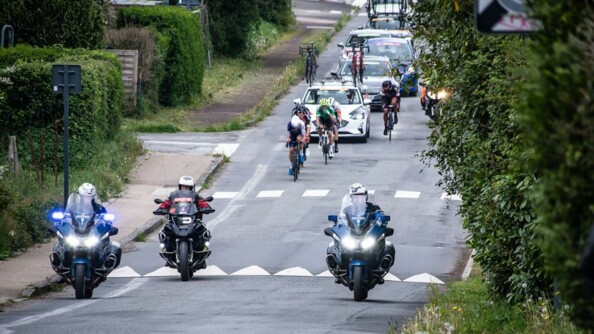 Deux motocyclistes de la garde républicaine sur une route de campagne, suivis d'un troisième motocycliste de la course. Derrière eux, quatre cyclistes et les véhicules de course.