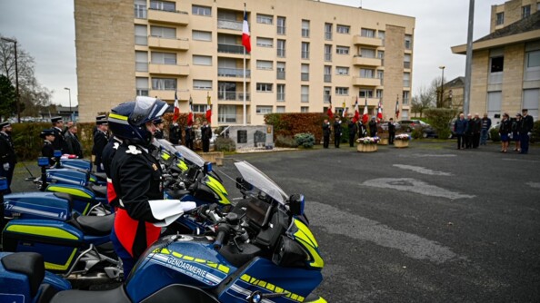 Au premier plan des motocyclistes de la gendarmerie de profil en grande tenue sur la place d'armes. En fond, de part et d'autre du mât des couleurs, une dizaine de porte-drapeaux.