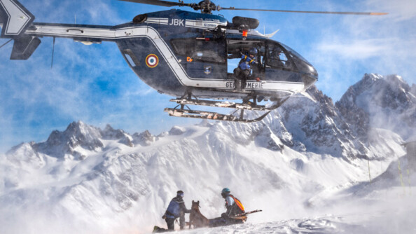 Deux gendarmes et un chien cyno en dessous d'un hélicoptère en pleine montagne.