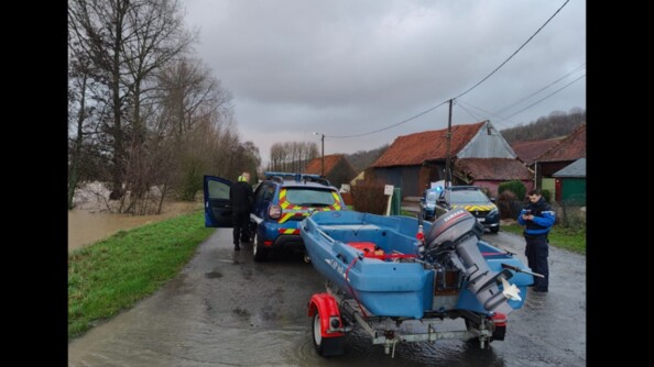 Deux véhicules de gendarmerie dont un tractant un bateau, sont positionnés au bord d'une route inondée. Sur l'image, se trouve un gendarme en tenue de service courant et un gendarme d'une brigade fluviale en tenue de plongeur.