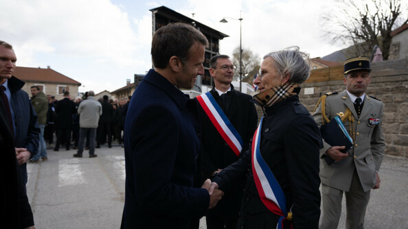Le président de la République, Emmanuel Macron, vêtu d'un costume sombre, serre la main d'une femme vêtue d'un trench noir, et portant sur le torse l'écharpe tricolore.
