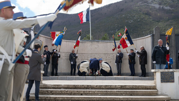 Devant un monument en pierre commémoratif, le président de la république, Emanuel Macron, se penche pour déposer au sol une gerbe de fleurs. Autour de lui se tiennent des hommes tenant un drapeau français.