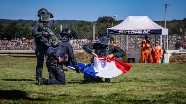 Trois militaires du GIGN au sol déploient un drapeau français. Au fond vers la droite, une tonnelle blanche siglée "LOHEAC". Au fond le public