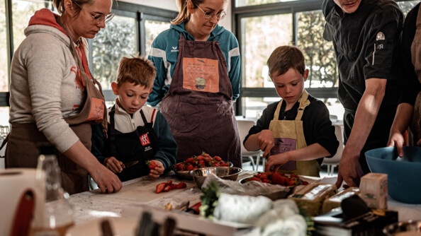 Les adultes encadrent la découpe de fraise fait par les enfants. Ils sont autour d'un plan de travail dans une cuisine.