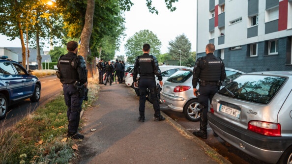 de dos, trois gendarmes en tenue noire et gilet pare-balles, derrière un attroupement et leur collègues, à droite des véhicules garés en bas de barres d'immeubles