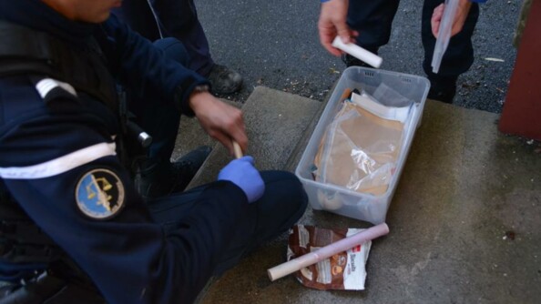 Un gendarme OPJ, de profil, dont on ne voit pas la tête, est accroupi devant une boîte en plastique transparent dans laquelle se trouve des papiers et divers produits liés au trafic de stupéfiants.