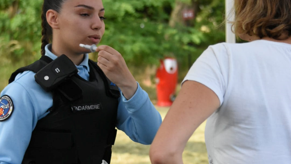 Une gendarme de face fait une démonstration de l'utilisation d'un éthylotest à une personne civile en tee-shirt blanc, vue de dos.