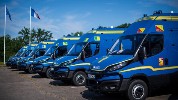 Six nouveaux véhicules de commandement et de transmission de la gendarmerie nationale, bleu avec des traits jaunes, stationnés en épis. Au fond les drapeaux francçais et européen.